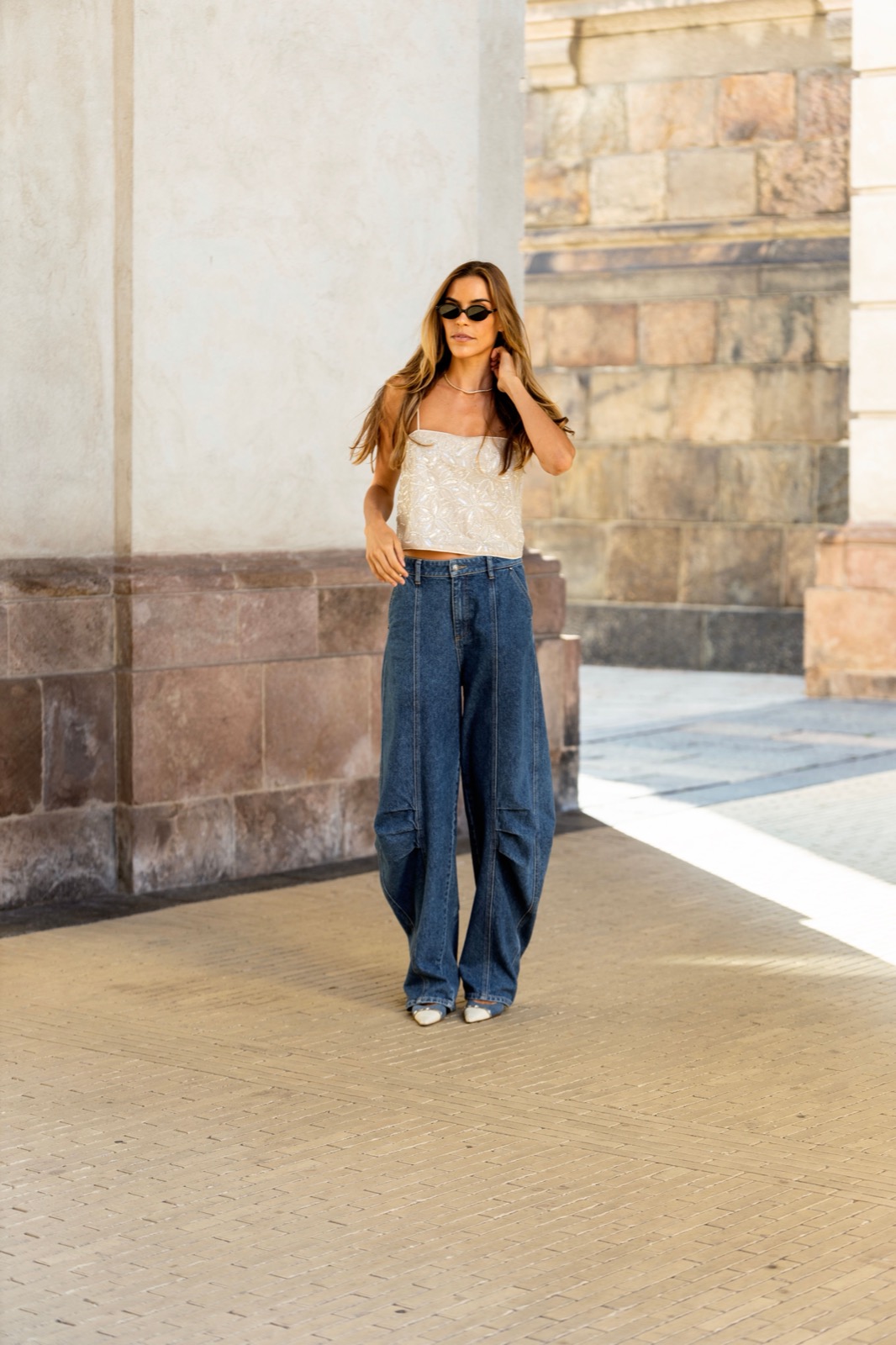 Woman in sunglasses, a pale tank top and wide-leg jeans standing by a stone building.